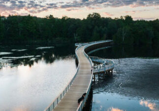 boardman lake loop trail drone 27 1440x542 Curved boardwalk over a serene lake at sunset.